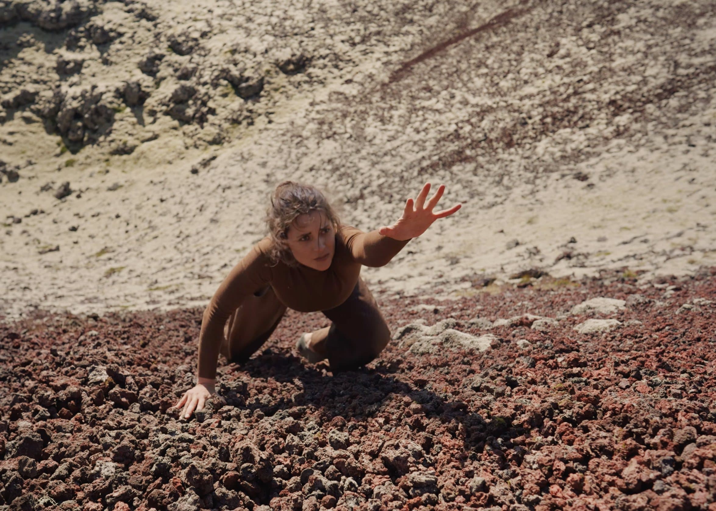 A dancer climbing a crater slope over volcanic rock, reaching forward.